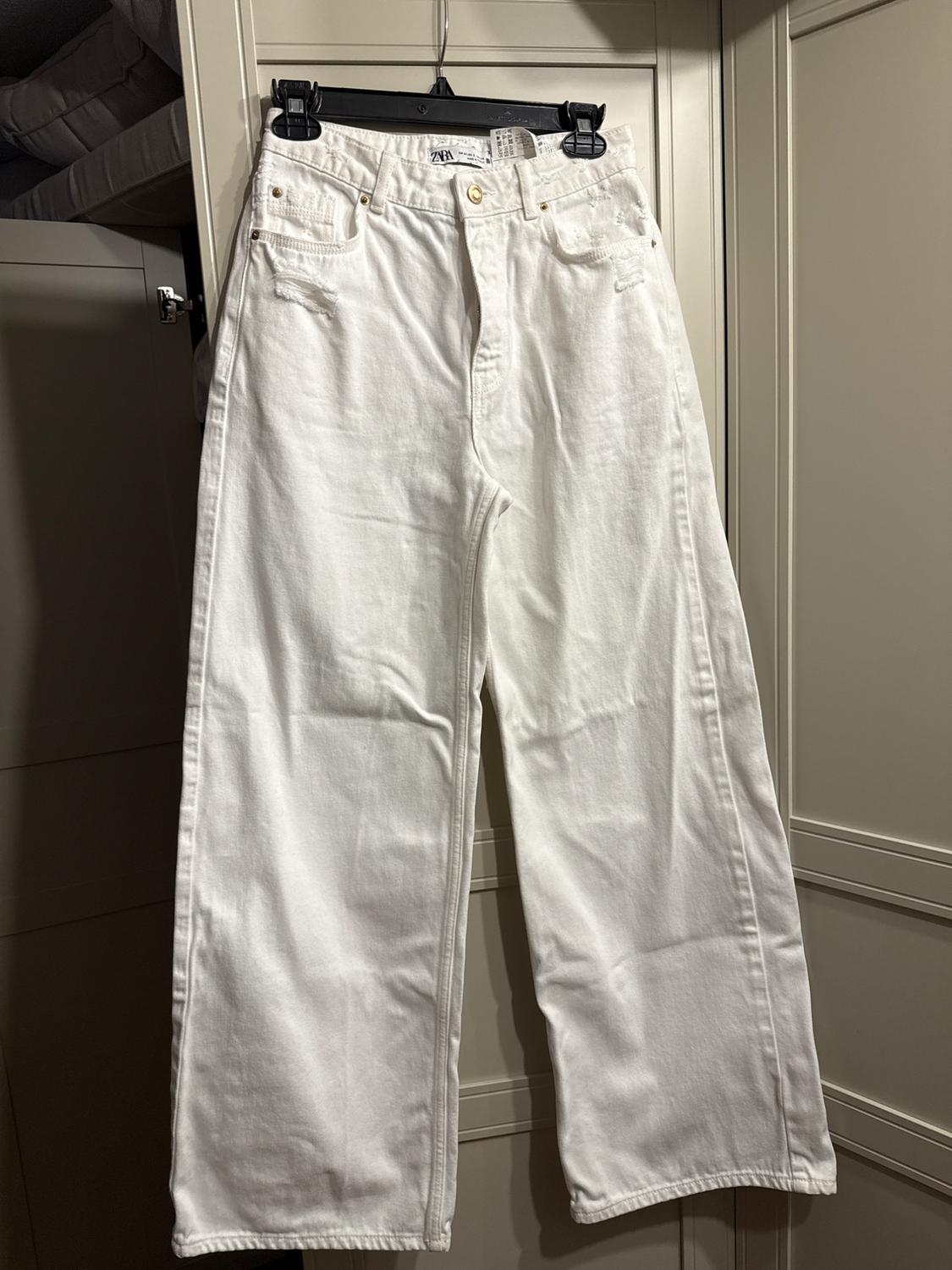 White denim pants hanging on a black hanger, with pockets and gold details, against a beige closet background.