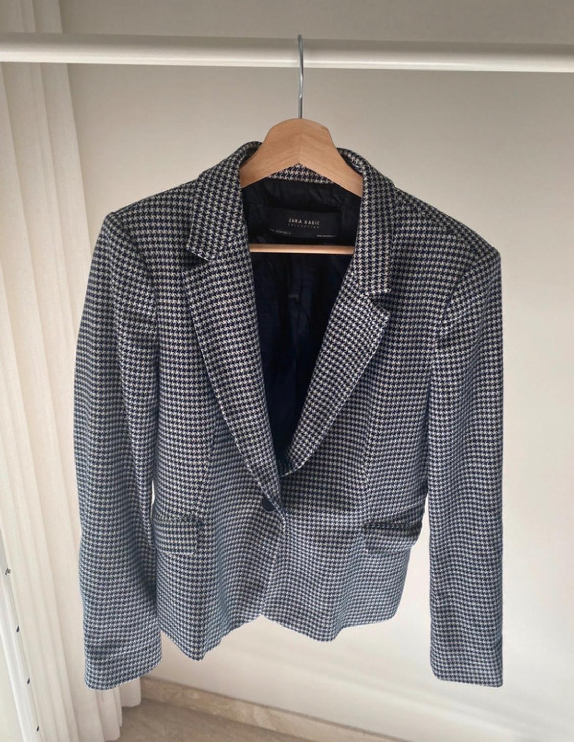 Black and white checked jacket hanging on a wooden hanger. Inside label reads "ZARA BASIC". Neutral background.