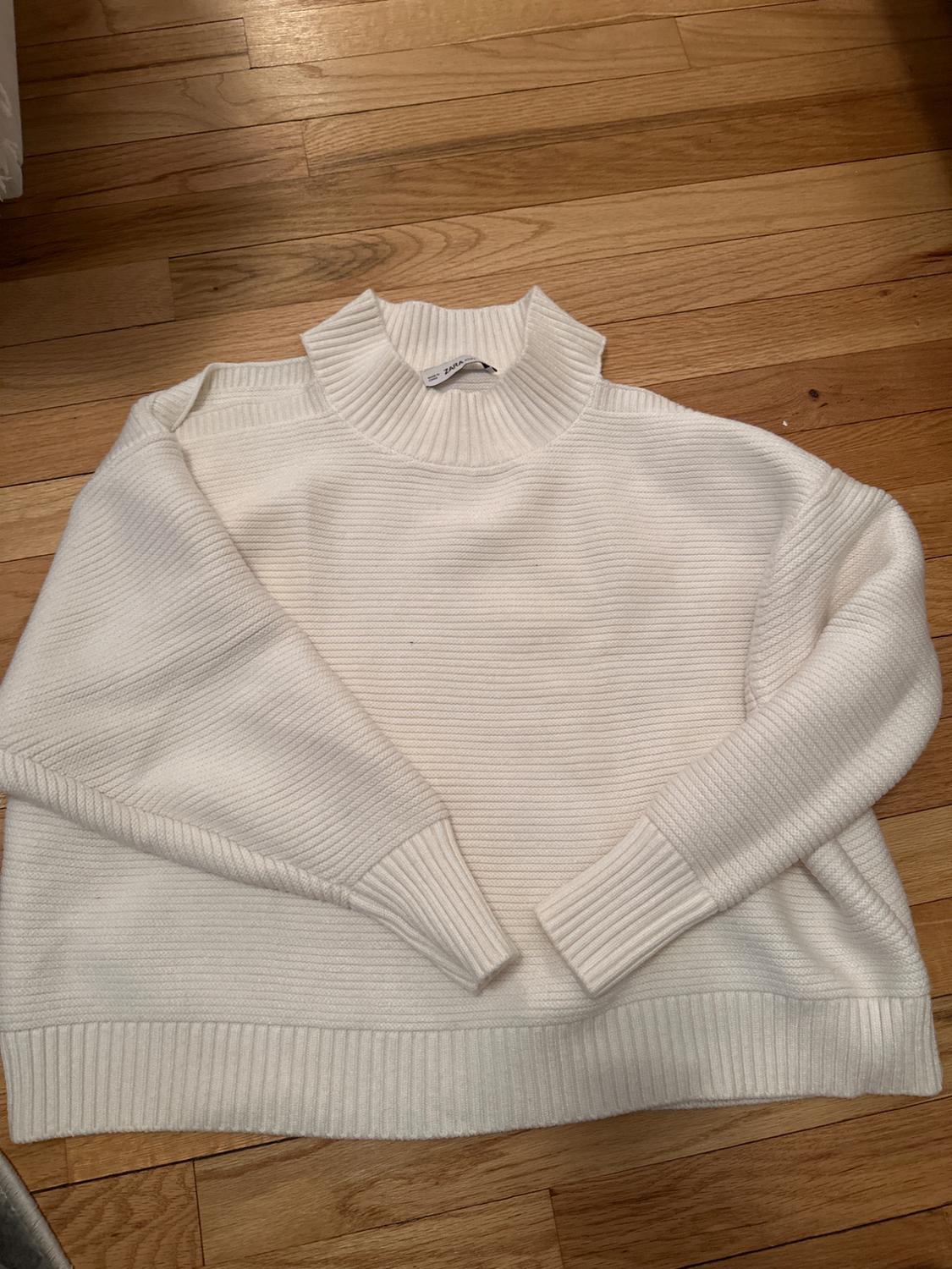 White chunky knit sweater with a high collar and long sleeves, placed on a light wooden floor. Visible tag on the collar.