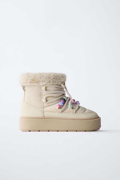 Seitenansicht eines cremeweißen Stiefelette mit bunten Perlen an den Schnürsenkeln und einem oberen Rand mit Fell-Effekt. Dicke beige Sohle.