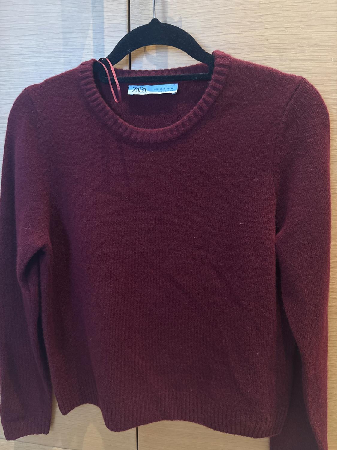 Burgundy long-sleeved jumper hanging on a black hanger, with a visible "ZARA" label, against a light wooden background.