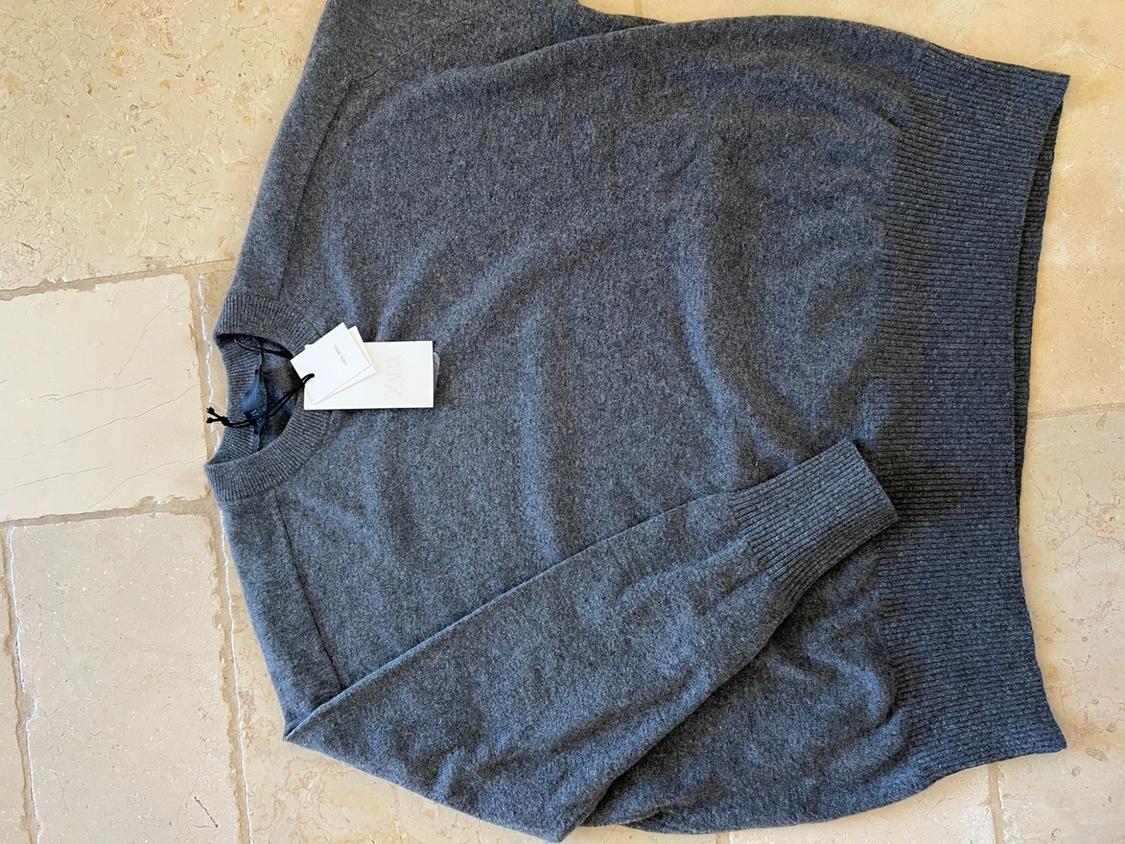 Grey knitted jumper laid out on a light-coloured tiled floor. It has a round neck and a white label with the text ZARA.