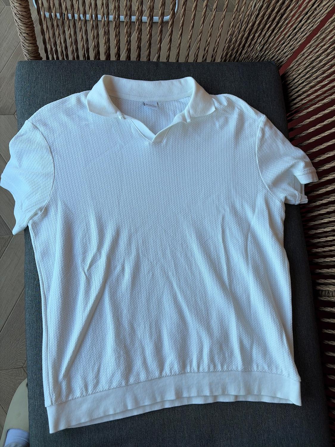 White short-sleeved polo shirt on a grey seat, with a slight texture and classic collar. Wicker chair and wooden floor in the background.