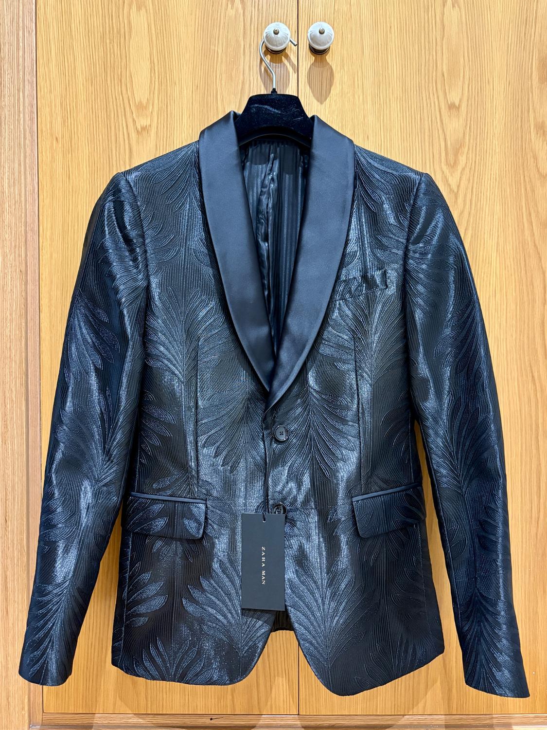 Black jacket with leaf texture and satin lapel, hanging on a wooden hanger. Visible tag: "ZARA MAN".