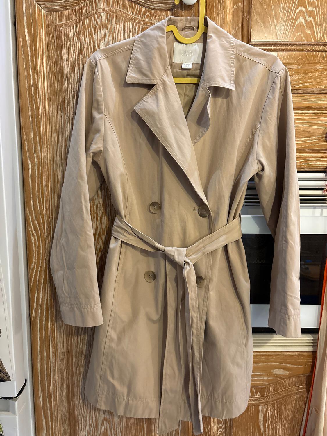 Beige trench coat hanging on a yellow hanger over a wooden door. It has a belt and front buttons. Visible label.