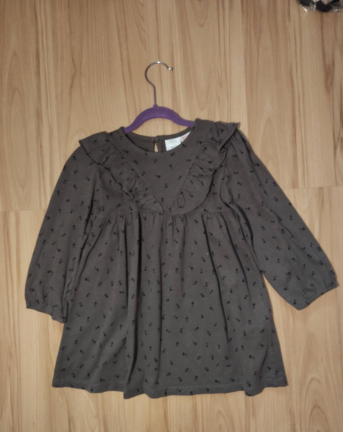 Dark grey dress with ruffles on the collar and long sleeves, hanging on a purple hanger over a light wooden floor.