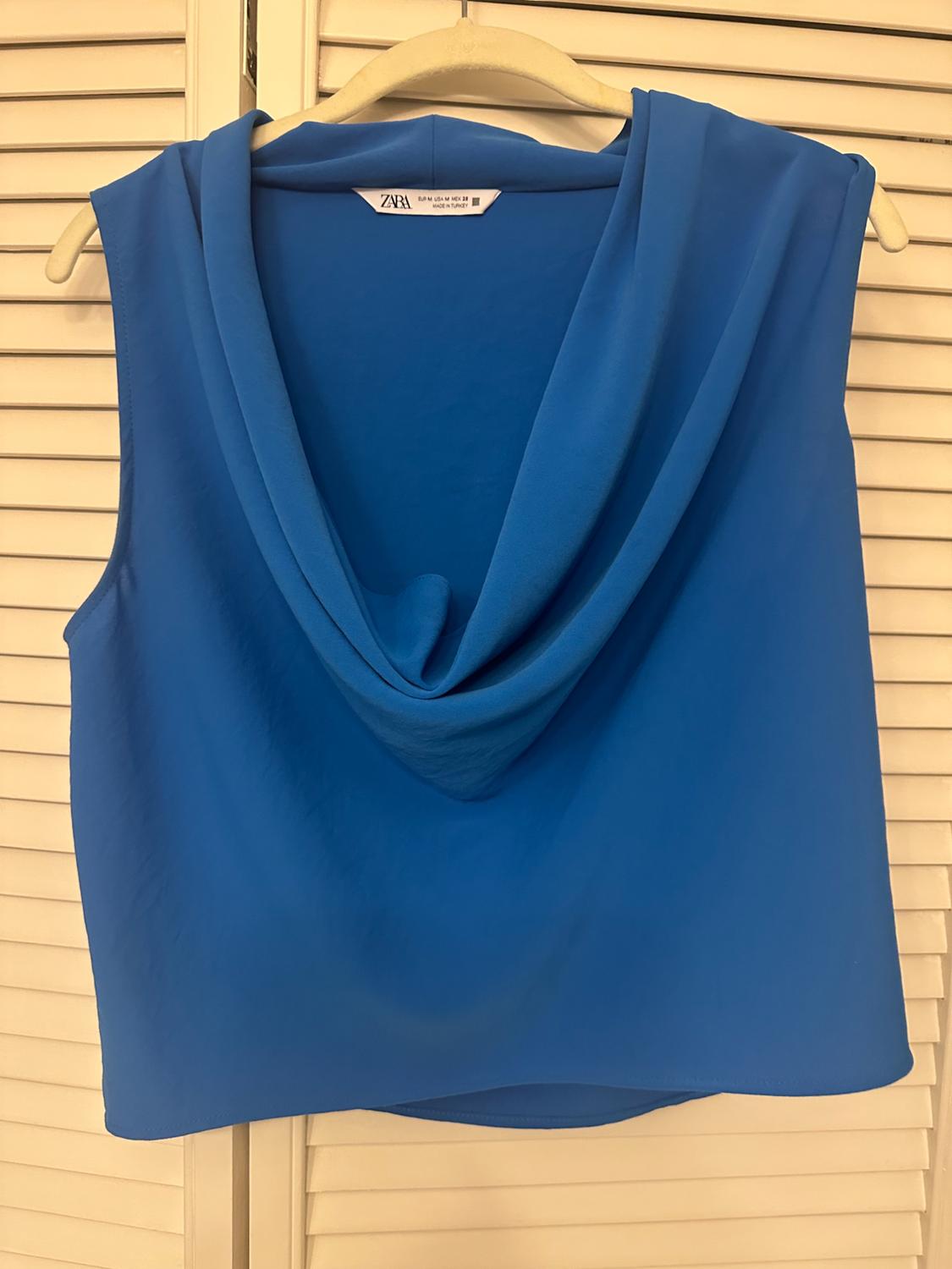 Blue sleeveless top with a draped neckline on a hanger over louvred doors. Text: ZARA EUR M USA M MEX 28 MADE IN TURKEY.