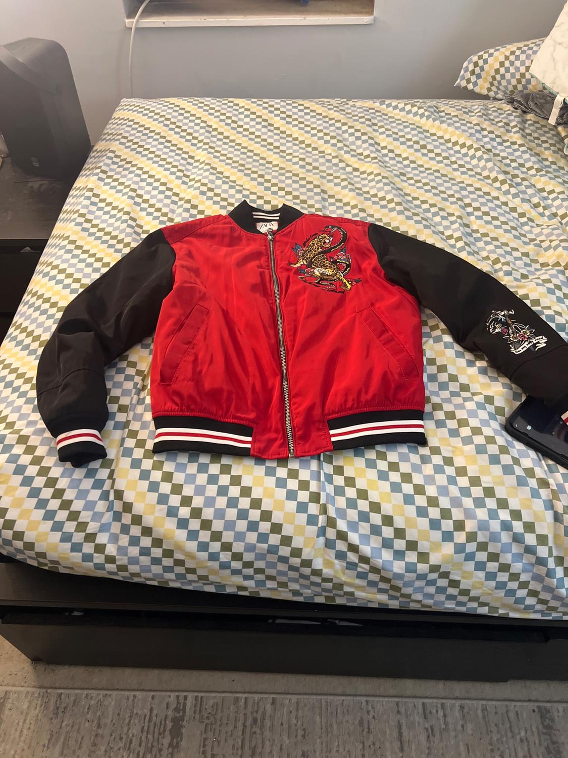Red bomber jacket with black sleeves, embroidery, and a ZARA label, laid out on a bed with plaid sheets.