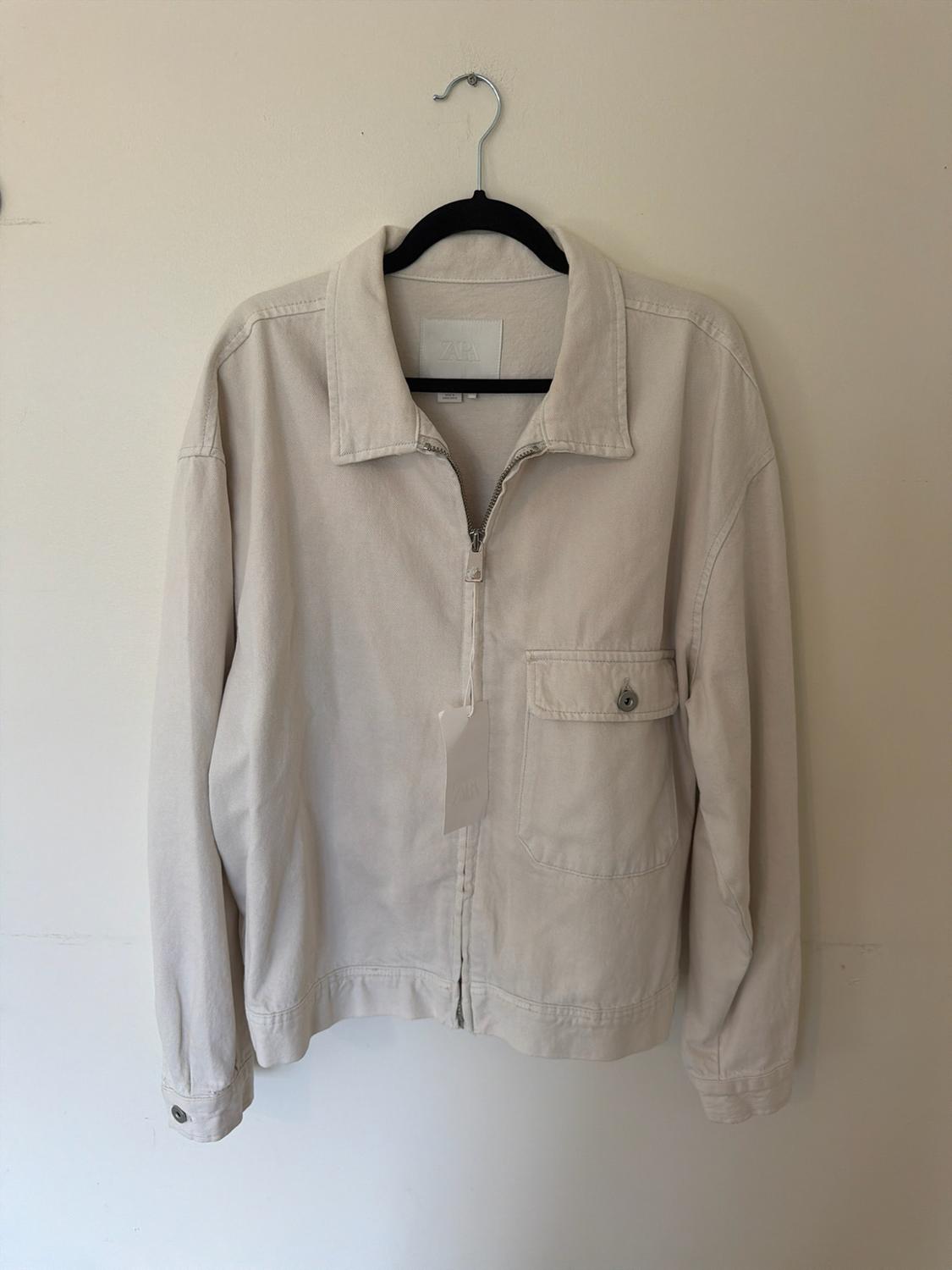 Beige zip-up jacket with collar and pocket, hanging on a black hanger against a white wall. Interior label with the text ZARA.