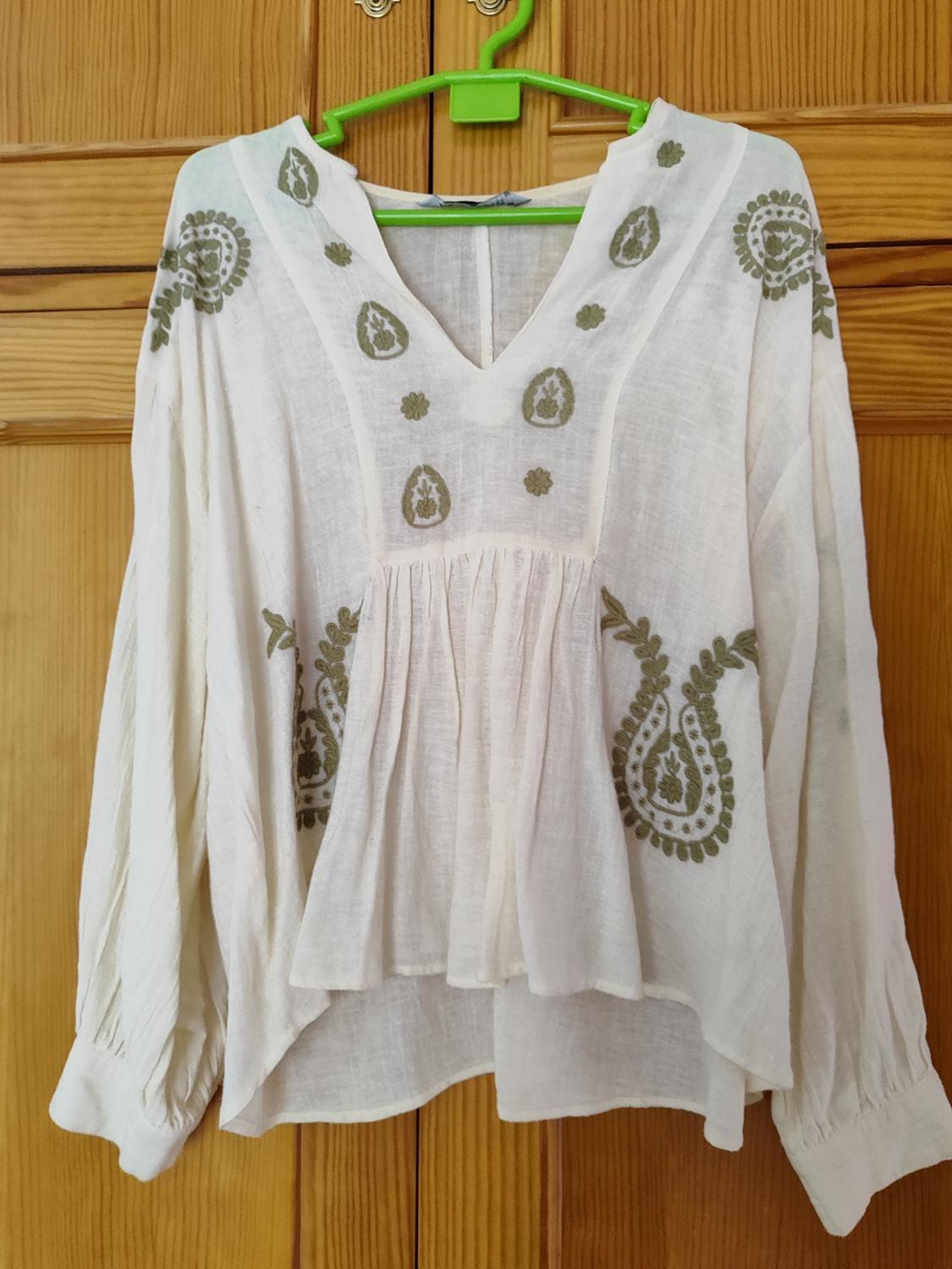 Long-sleeved white blouse with green floral embroidery, hanging on a green hanger against a wooden background.