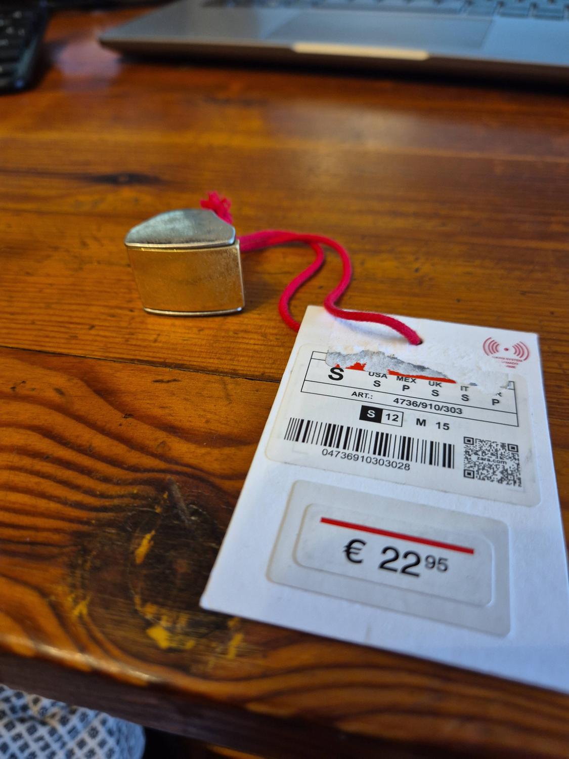 White tag with a pink cord and metal piece on a wooden table. Text: ART.: 4736/910/303, €22.95.