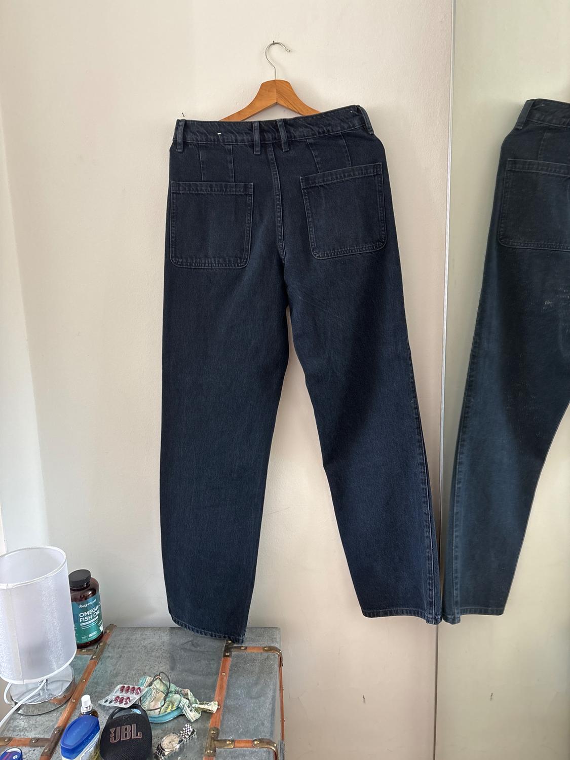 Dark jeans on a hanger against a white wall next to a mirror. Below, a trunk with a lamp, a JBL speaker and a bottle of OMEGA FISH OIL.