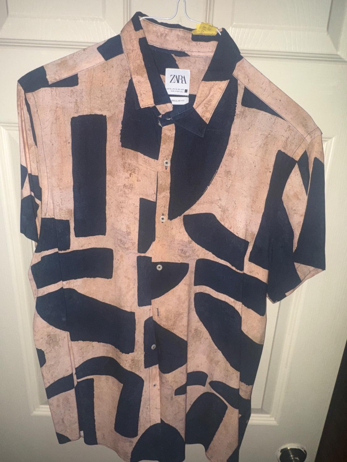 Short-sleeved shirt with an abstract print in beige and black tones, hanging on a door. Visible label: "ZARA".