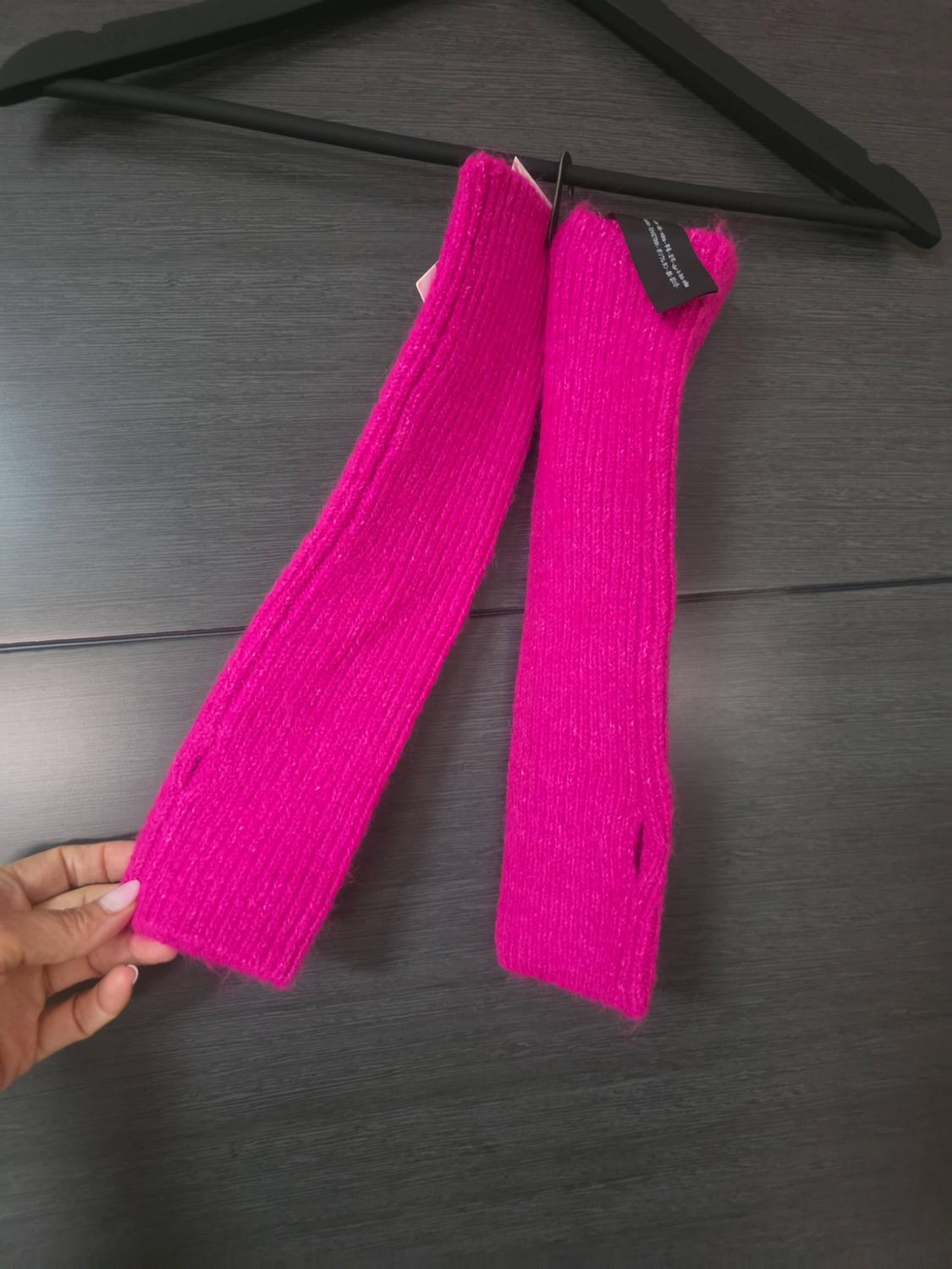Fuchsia pink knitted leg warmers hang from a black coat hanger against a dark grey background. A hand grips the end of one of them.