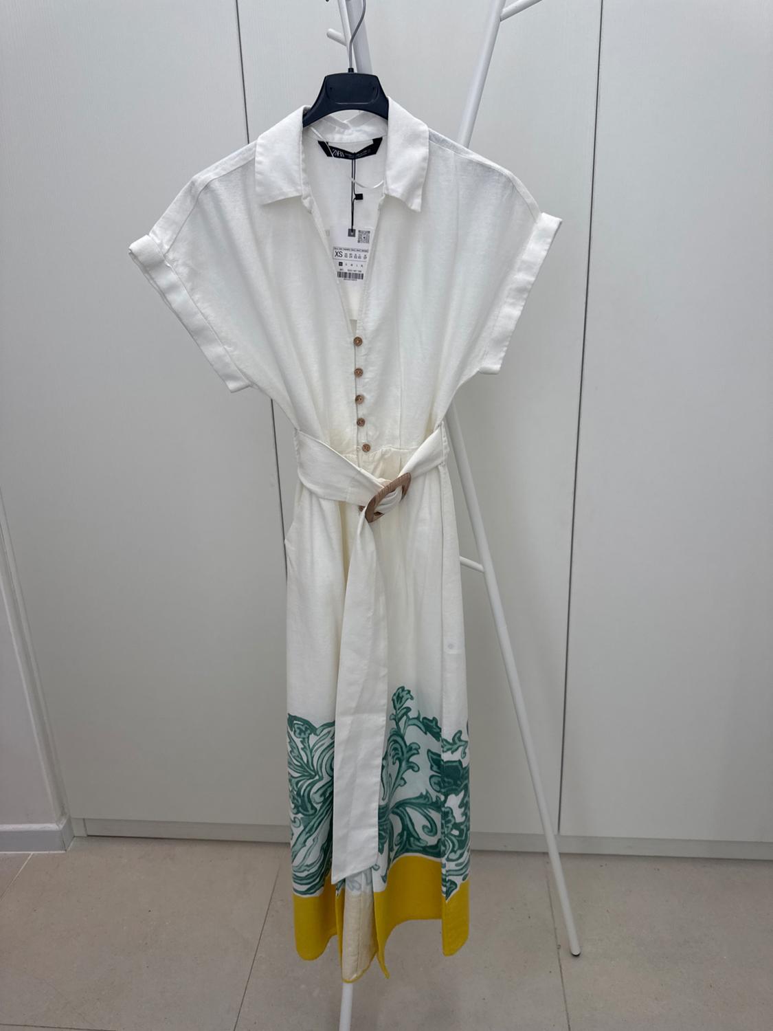 White dress with brown buttons, belt and green and yellow print on the hem, hanging on a white hanger.