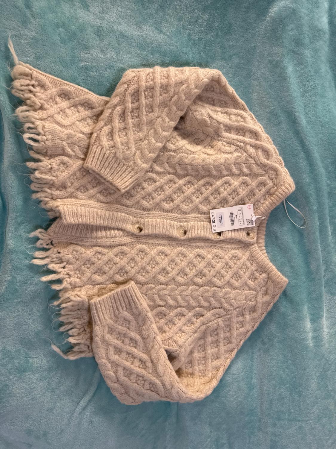 Beige knitted cardigan with braided design and fringing on the edges, on a blue background. Visible label with price and size.