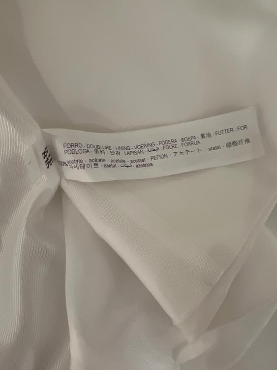 White fabric label with multi-language text indicating "LINING" and "100% acetate". Wrinkled white fabric background.