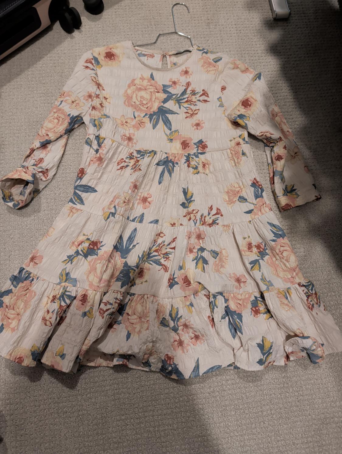 Long-sleeved dress with a floral print in pastel shades on a beige background, hanging on a hanger over a grey carpet.