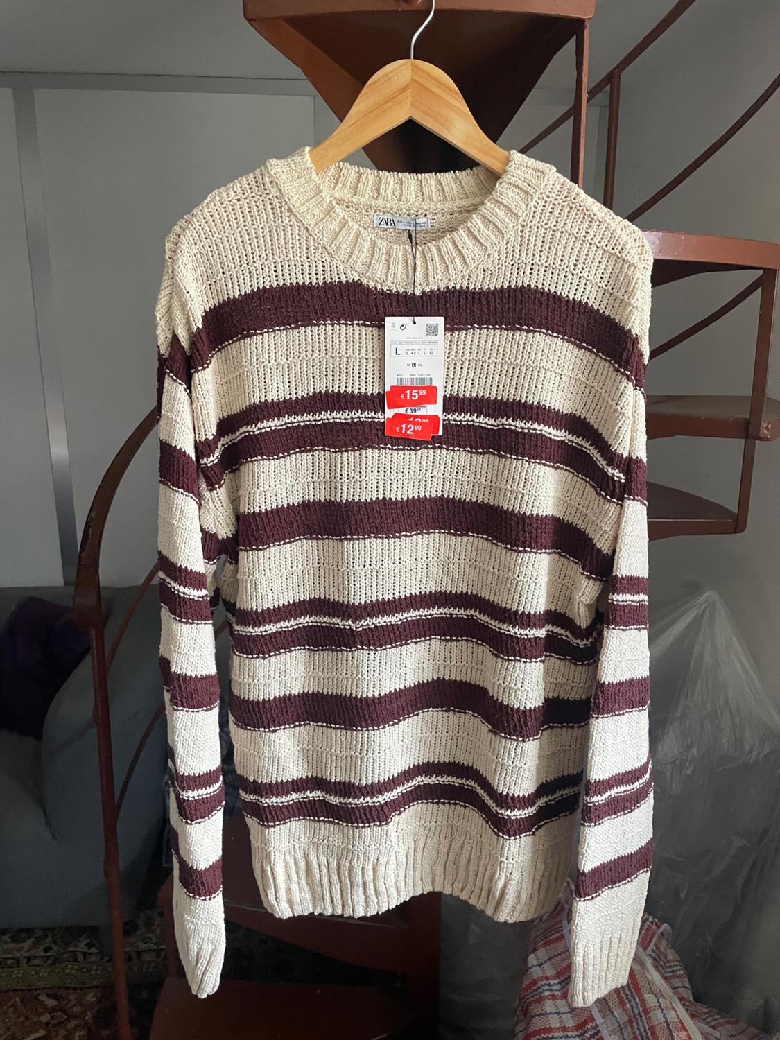 Knitted jumper with brown and beige stripes hanging on a wooden hanger. Discounted price tag visible. Staircase in the background.