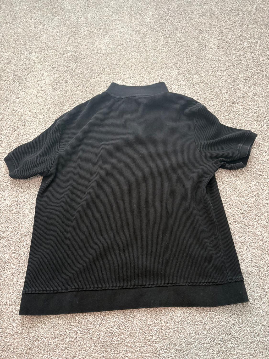 Black short-sleeved T-shirt seen from the back, on a beige rug. Fabric with ribbed texture and classic collar.