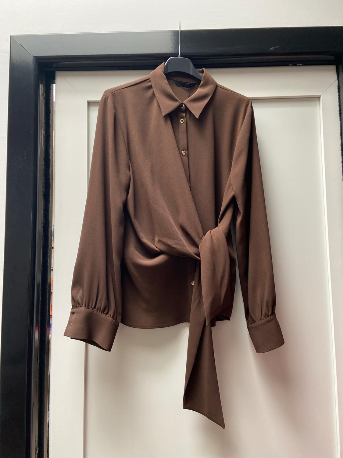 Brown long-sleeved collared blouse with buttons and a waist tie, hanging from a black coat hanger on a white door.