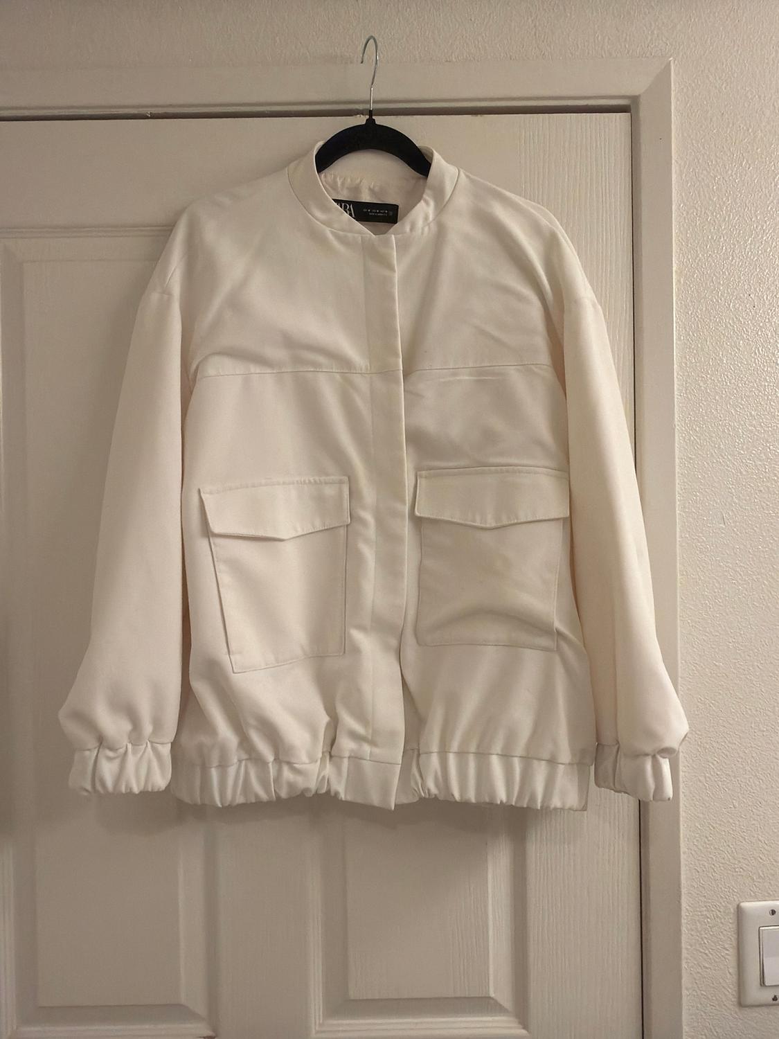 White jacket hanging on a hanger over a door. It has large pockets and a minimalist design with a round neck.