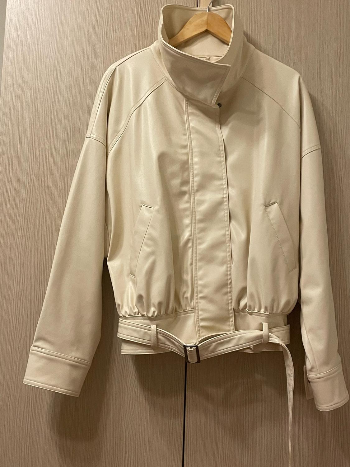 Cream high-necked jacket with side pockets and a belt at the hem, hanging on a hanger against a light wood background.