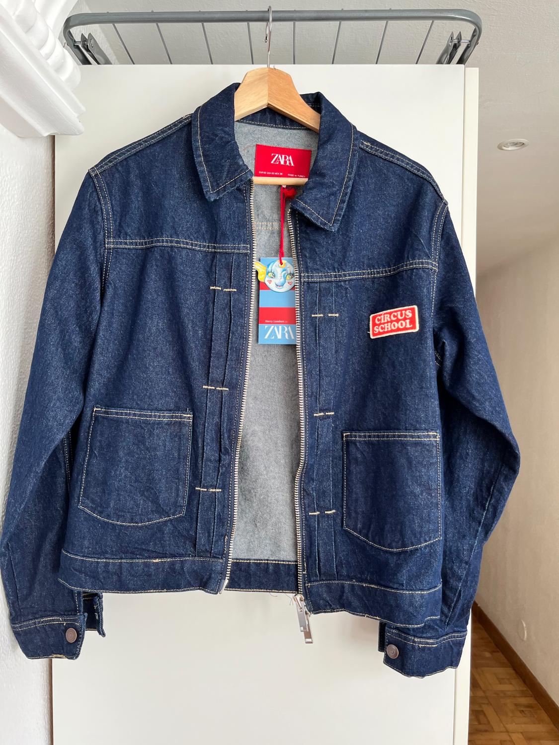 Blue denim jacket hanging on a wooden coat hanger. Red "ZARA" label and "CIRCUS SCHOOL" patch on the chest.