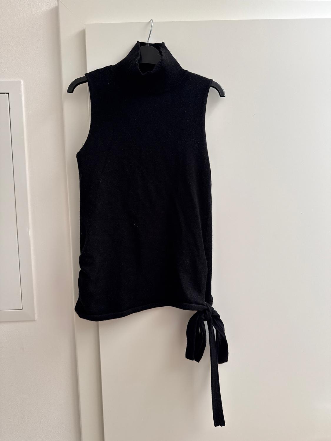 Black sleeveless high-neck top with a tie on the side, hanging on a hanger over a white door.