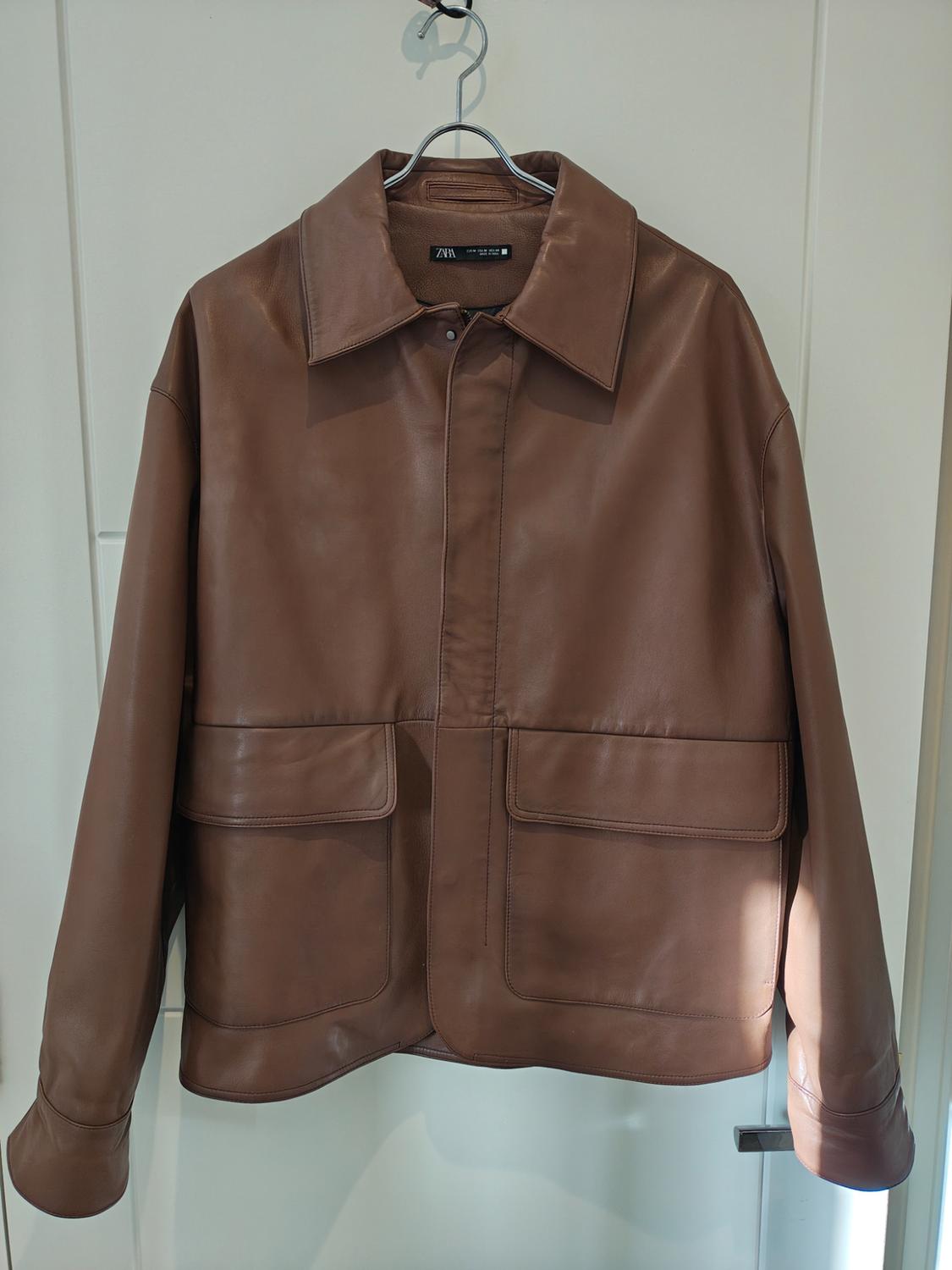 Brown leather jacket hanging on a hanger, with large pockets and a classic collar. Visible label with text "ZARA".