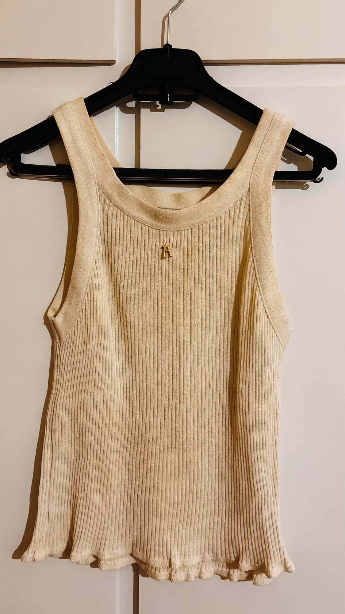 Beige ribbed sleeveless top hanging on a black hanger, with a small gold embellishment in the centre of the neckline.