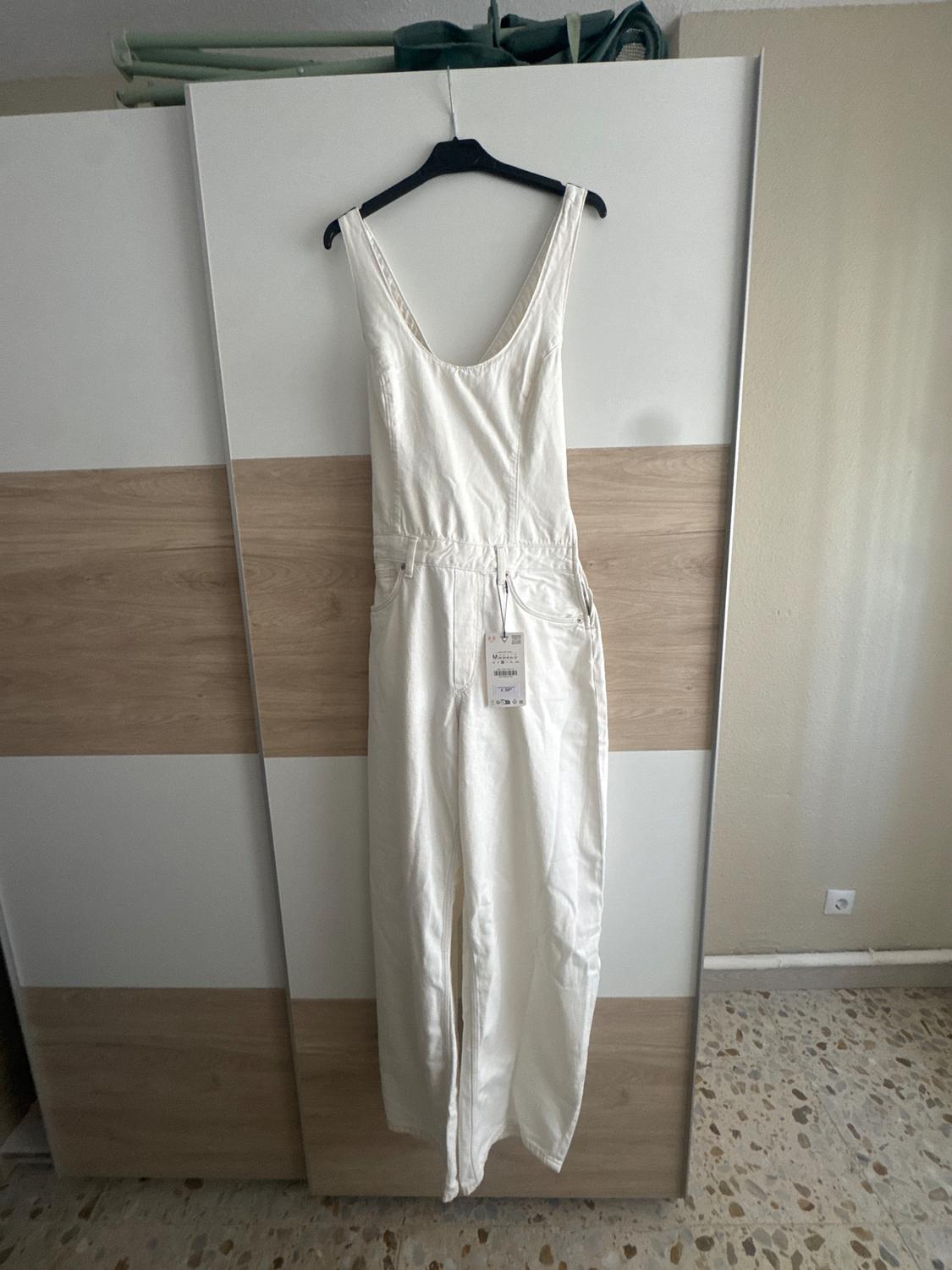 White sleeveless jumpsuit hanging on a black hanger in front of a wooden wardrobe. Label visible in the centre of the jumpsuit.