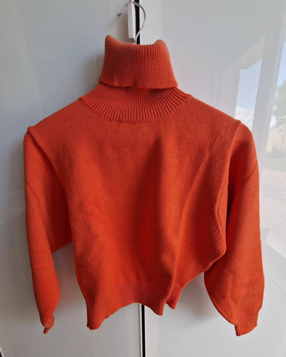 Orange knitted jumper with a high neck, hanging on a hanger in front of a white wardrobe.