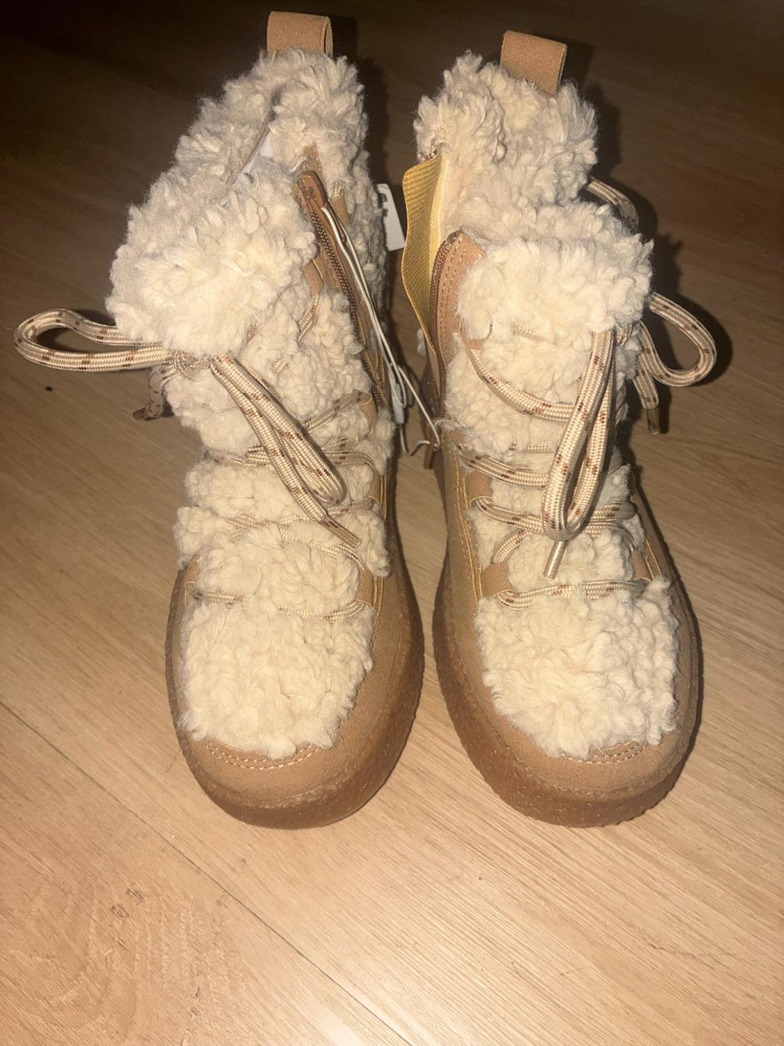 Camel-colored winter boots with plenty of faux fur, laces, side zipper, and a thick sole, on a wooden floor.
