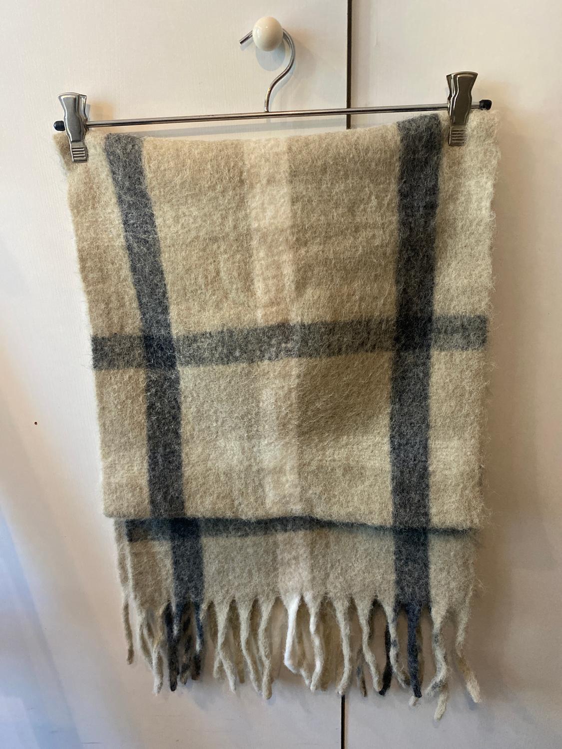 Beige wool scarf with grey checks and tassels hanging on a metal hanger over a white door.