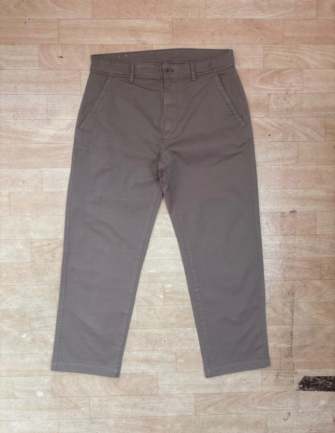 Casual brown trousers with side pockets and button fastening, laid out on a light wooden floor.