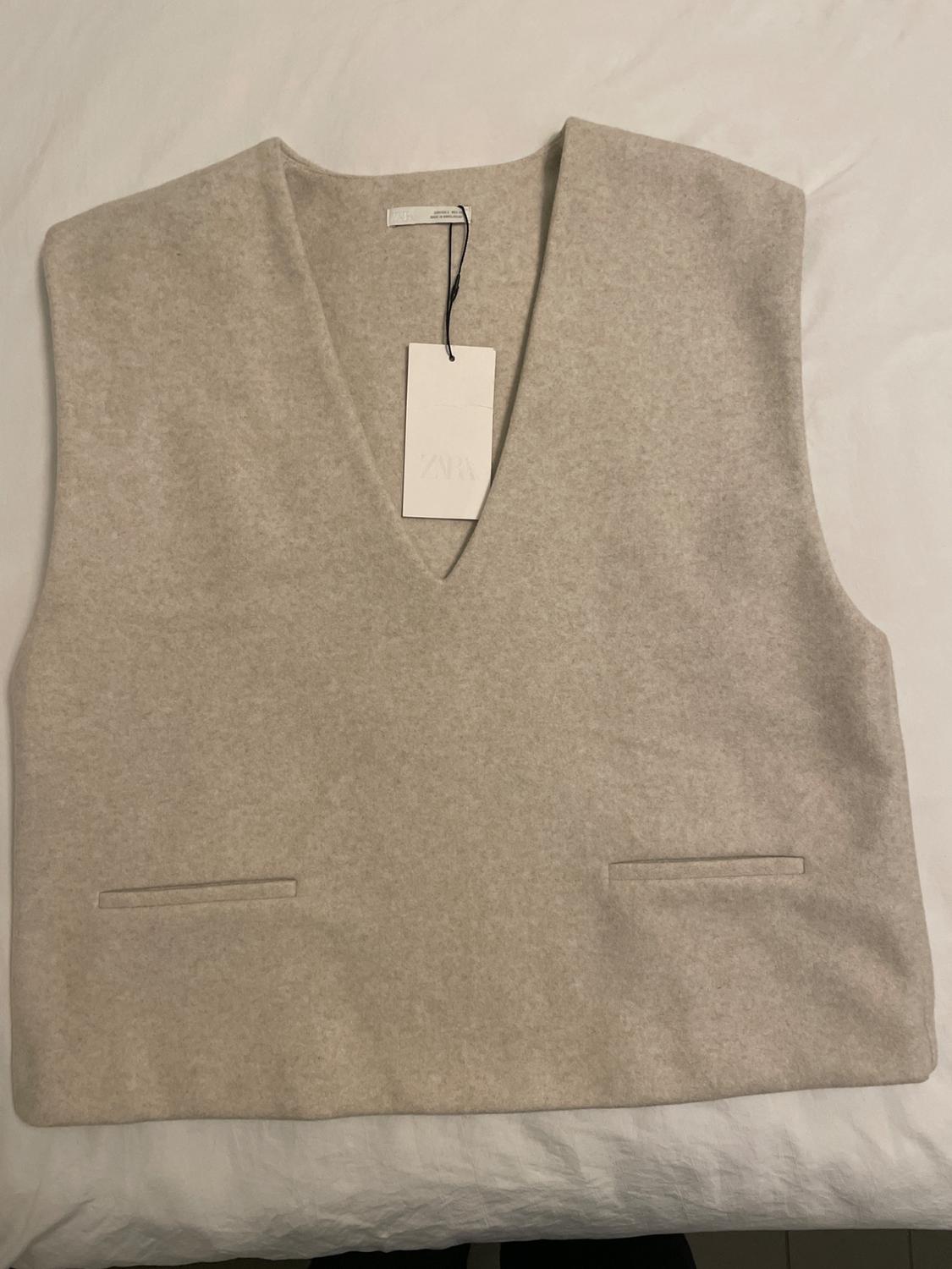 Beige V-neck sleeveless waistcoat on a white surface. White swing tag with "ZARA" text.