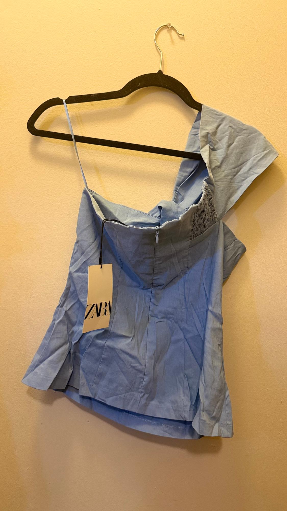 Light blue camisole with thin straps and "ZARA" tag, hanging on a black hanger against a beige wall.