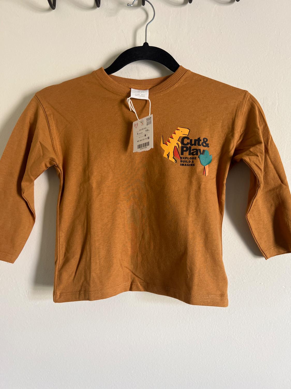 Children's long-sleeved brown T-shirt with dinosaur design and "Cut & Play, Explore, Build & Imagine" text, hanging on a black hanger.