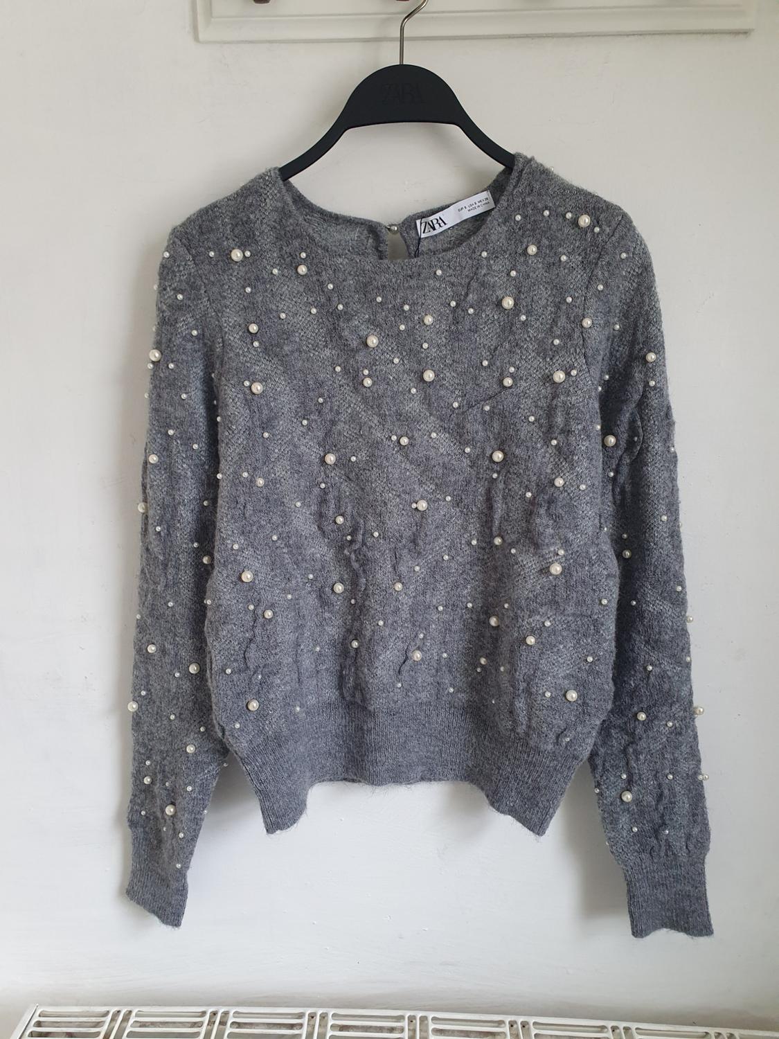 Grey jumper with decorative pearls hanging on a black hanger with the label visible. White background.