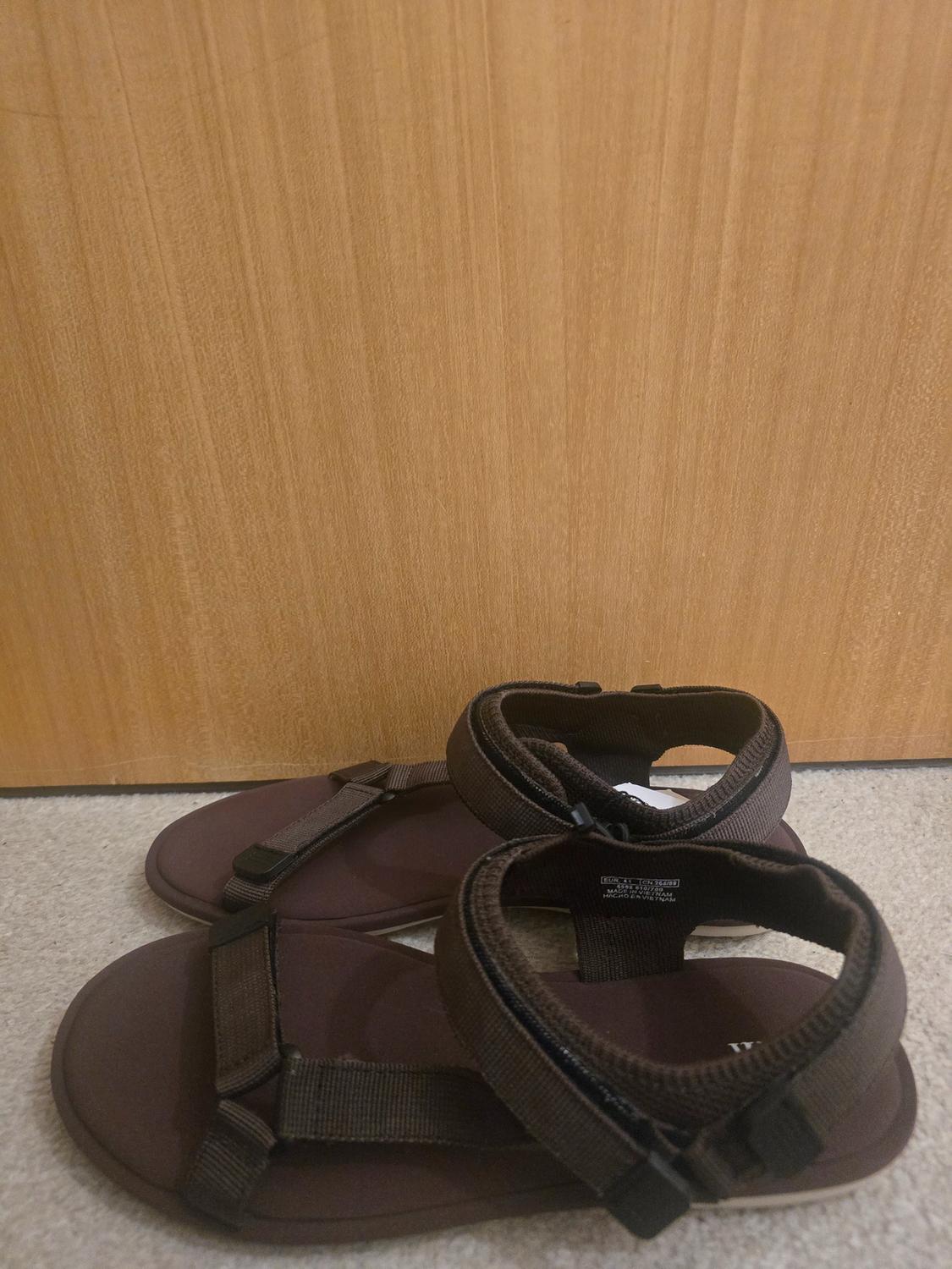 Brown sandals with adjustable straps on a beige rug and light wood background. Visible text on the insole.