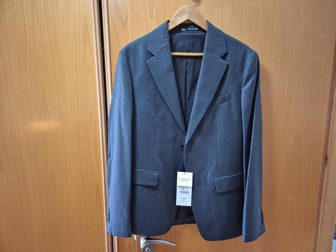 Grey jacket hanging on a wooden hanger over a light wooden door. Visible label with text "Zara".
