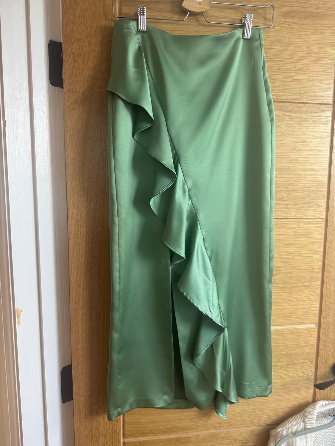 Green satin flounced skirt hanging on a wooden hanger over a light wooden door.