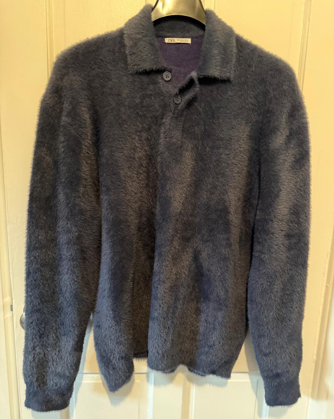Dark grey fleece jumper hanging on a hanger, with collar and buttons visible. White door background. Label visible.