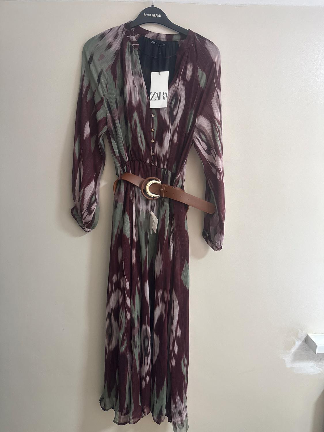 Long, printed dress in shades of brown and green, with a brown belt and gold buckle, hanging on a black River Island hanger.