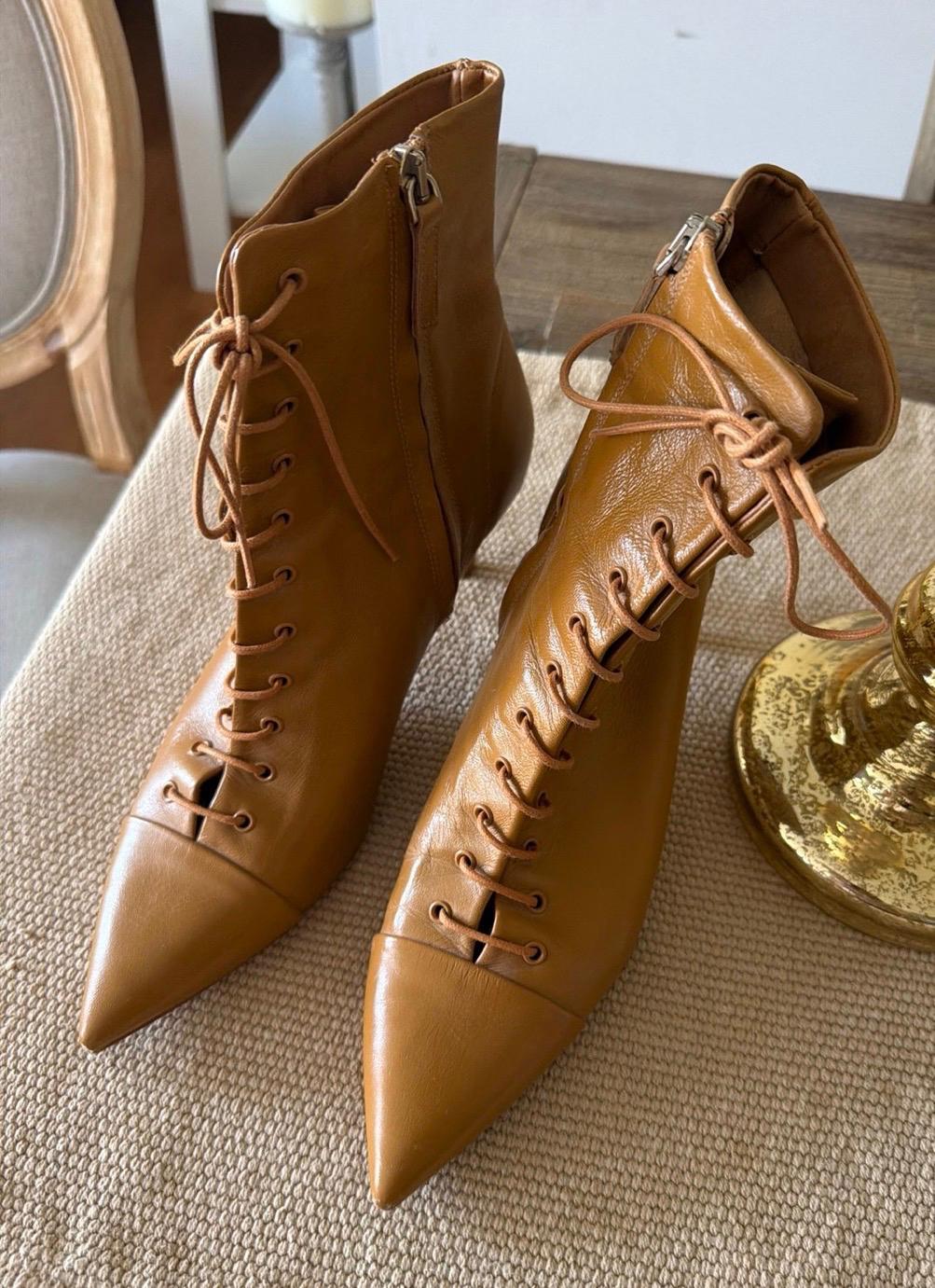 Brown leather ankle boots with laces and pointed toes, displayed on a beige woven surface beside a gold detail.