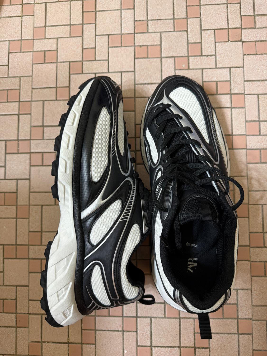 Black and white trainers on a tiled floor. One profile view and one from above. Inner text: Zara Ortholite.