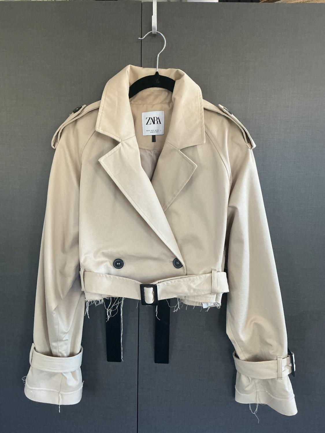 Short beige trench coat hanging on a hanger against a dark background. It features a frayed hem, a belt, and a label with the text ZARA.
