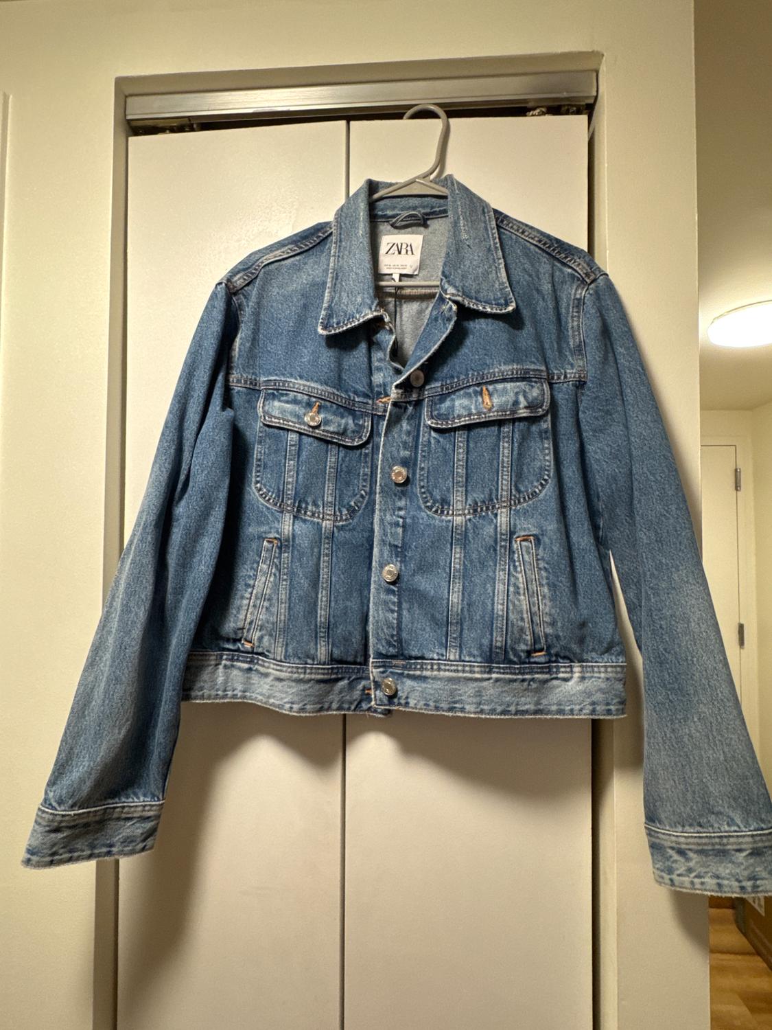 Blue denim jacket hanging on a hanger in front of a white door. Visible tag with text "ZARA".