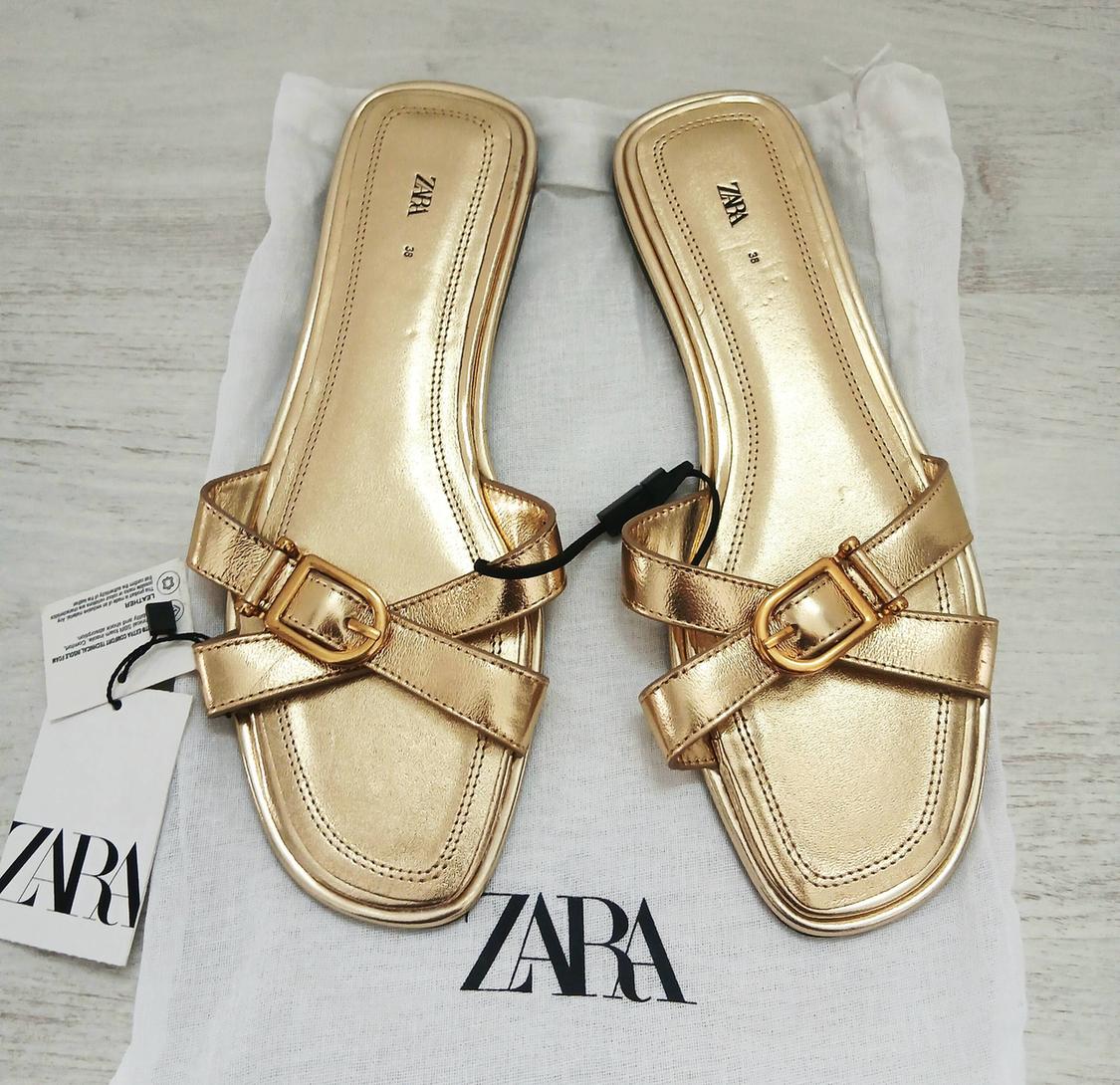Gold sandals with buckles on a white bag with the Zara logo. Visible hang tag on the left.